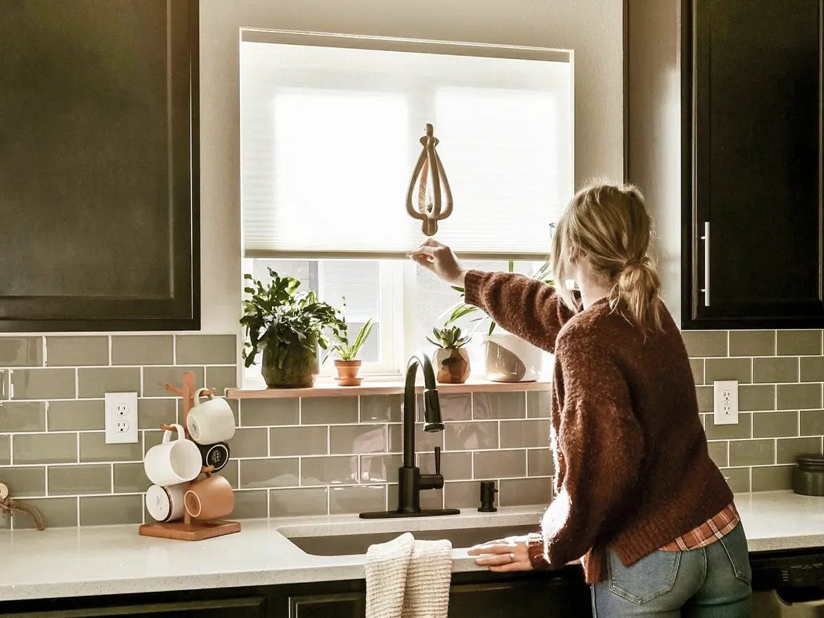 Woman adjusting cellular shades above a kitchen sink with plants on the windowsill.