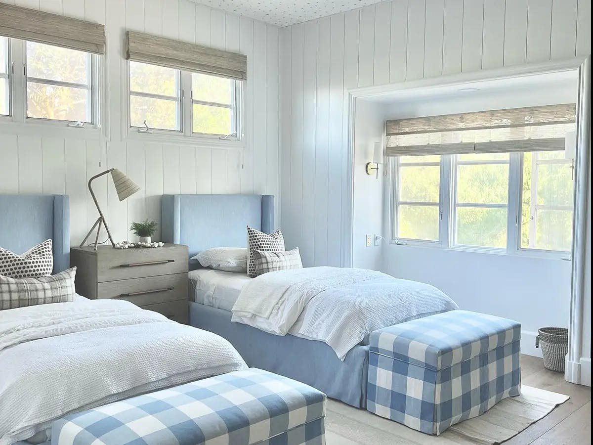 Bedroom with two twin-sized beds. The headboards are blue and the sheets are white. At the foot of each bed are blue gingham benches. On the windows are light colored woven wood shades.