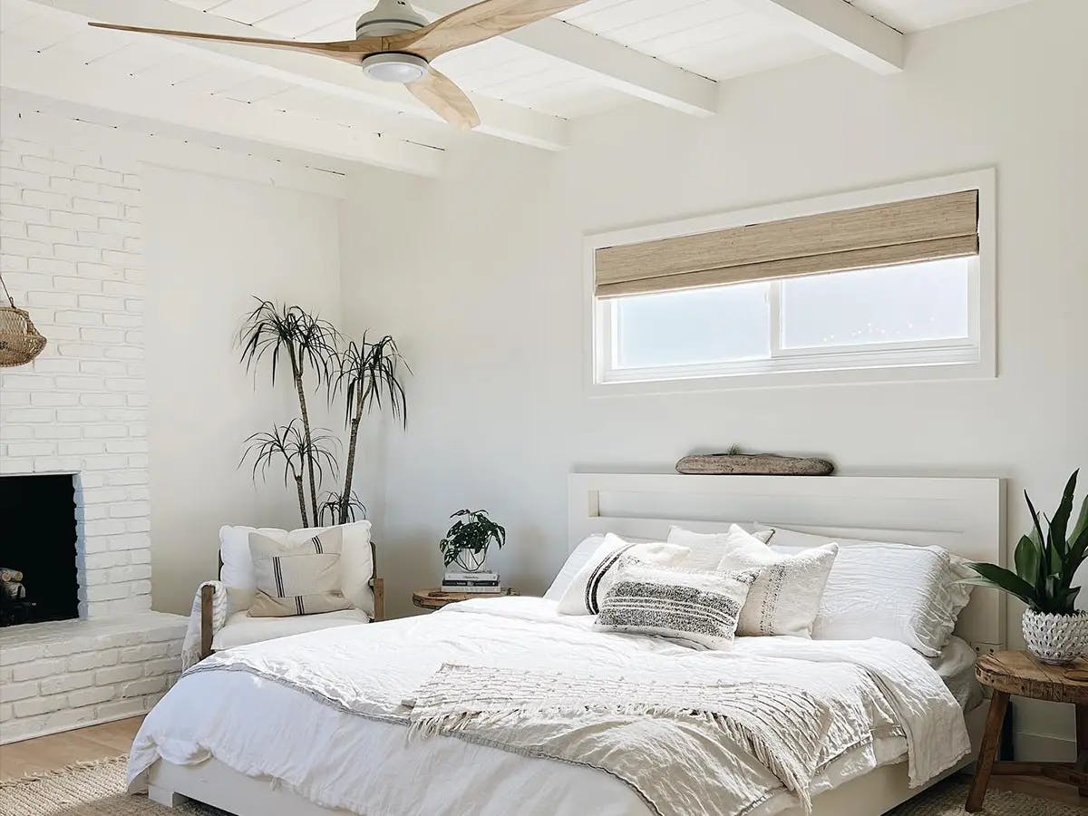 Bedroom with white walls and white ceilings. The bed is large and has white bedding. Above the bed is a window with woven wood shades.