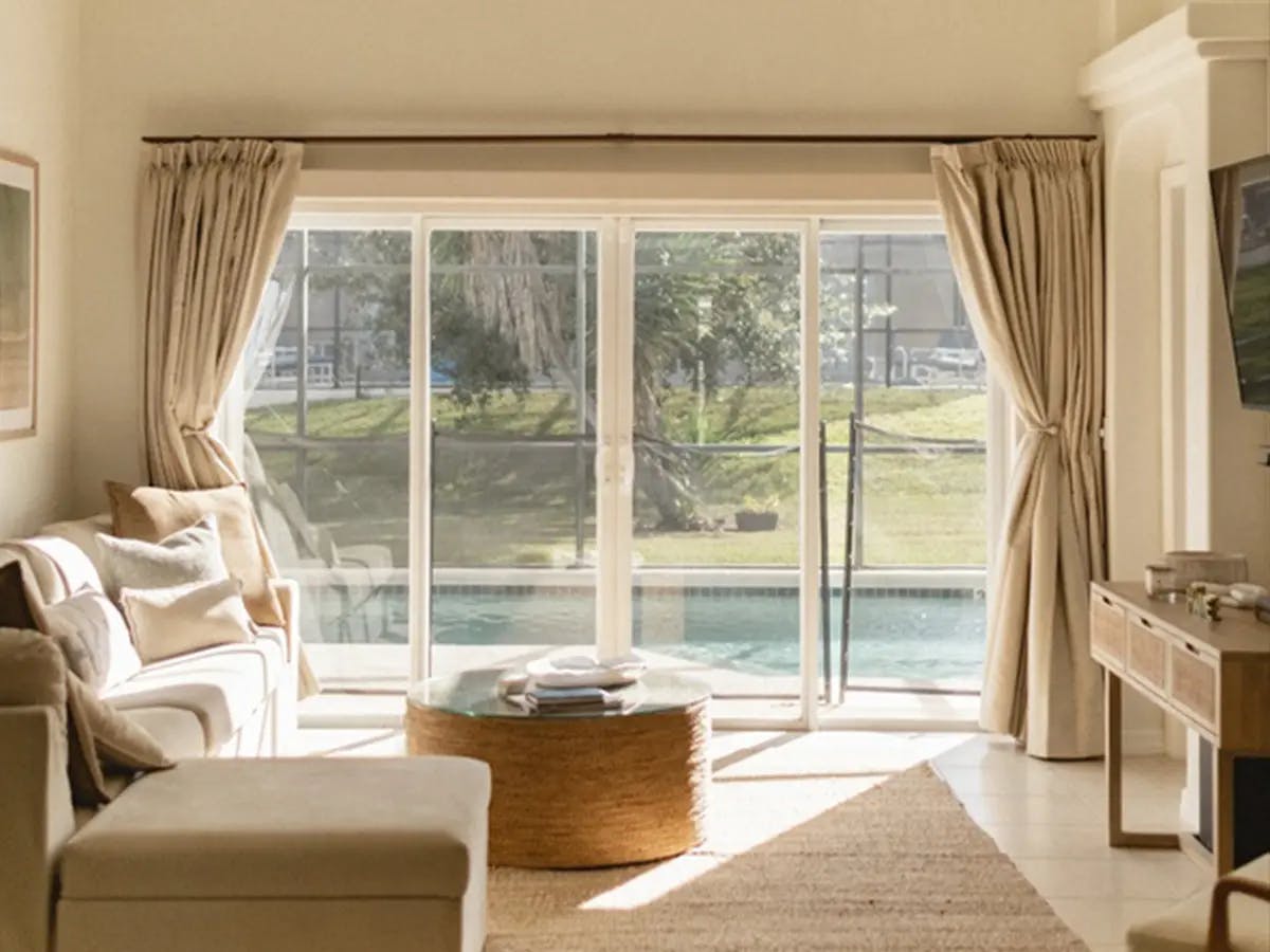 Living room with a large window looking out into a backyard with a pool. The furniture has neutral and warm tones. The window is framed by neutral colored floor-length drapes.