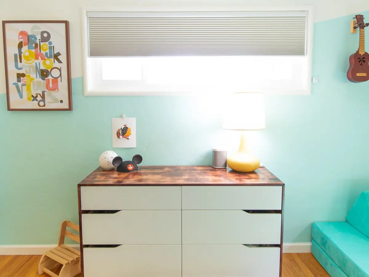A dresser with a lamp, framed art and shades on the window.