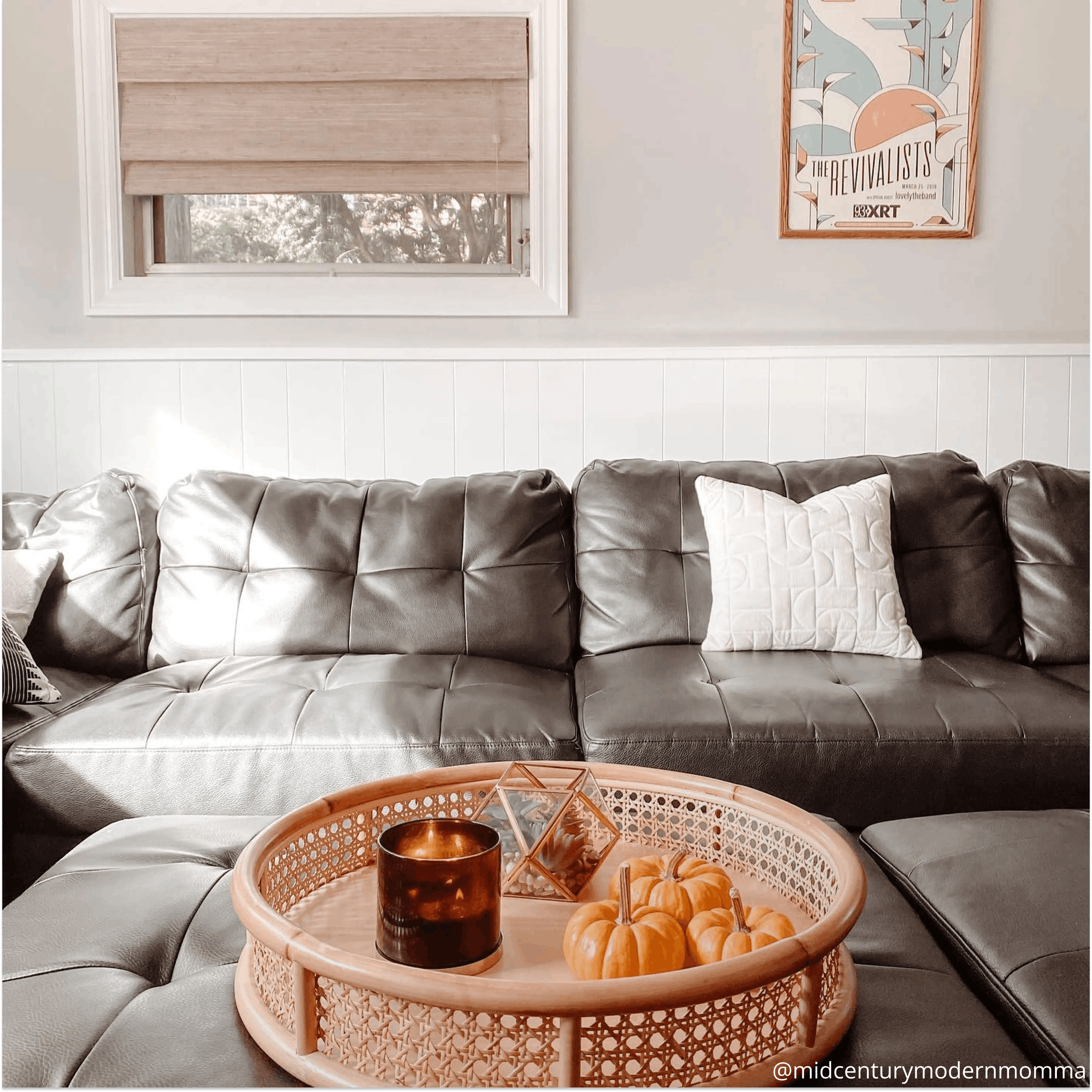 Black leather couch with a white throw pillow. The ottoman features a wicker tray with mini pumpkins, mini terrarium and candle. The window above the couch is covered with a neutral-colored Roman shade.