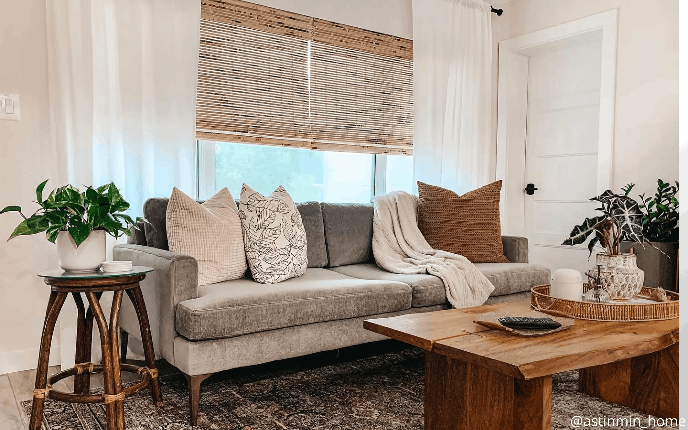 Gray velvet couch with various throw pillows in orange gingham, brown and leaf patterns. It also features a gray throw blanket. The window behind the couch features woven wood shades layered with sheer white curtains.