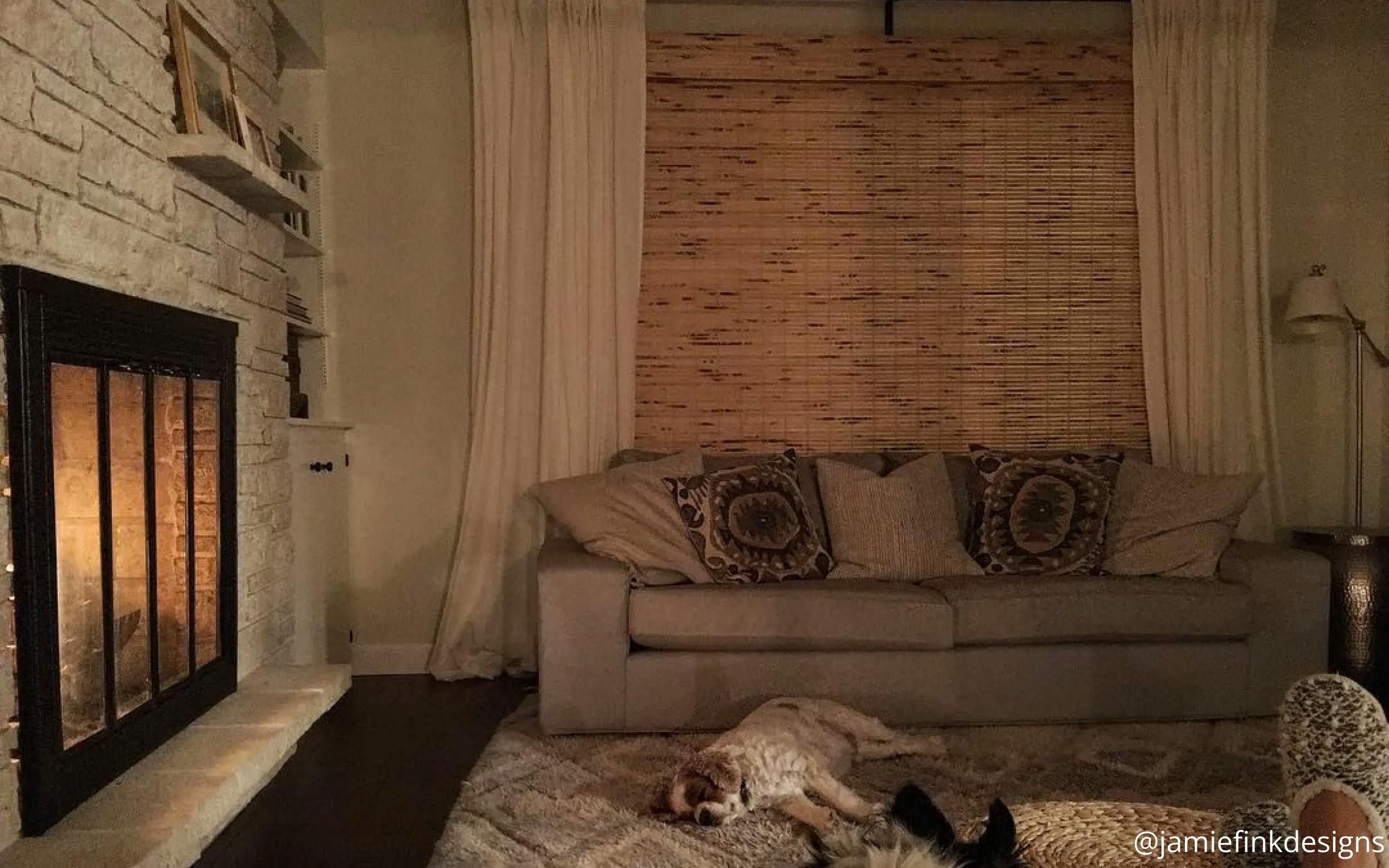 Dark living room space lit by the fireplace. The window is fully covered by woven wood shades layered under white curtains. Above the fireplace is a mantle with picture frames. A white dog sleeps on the rug.