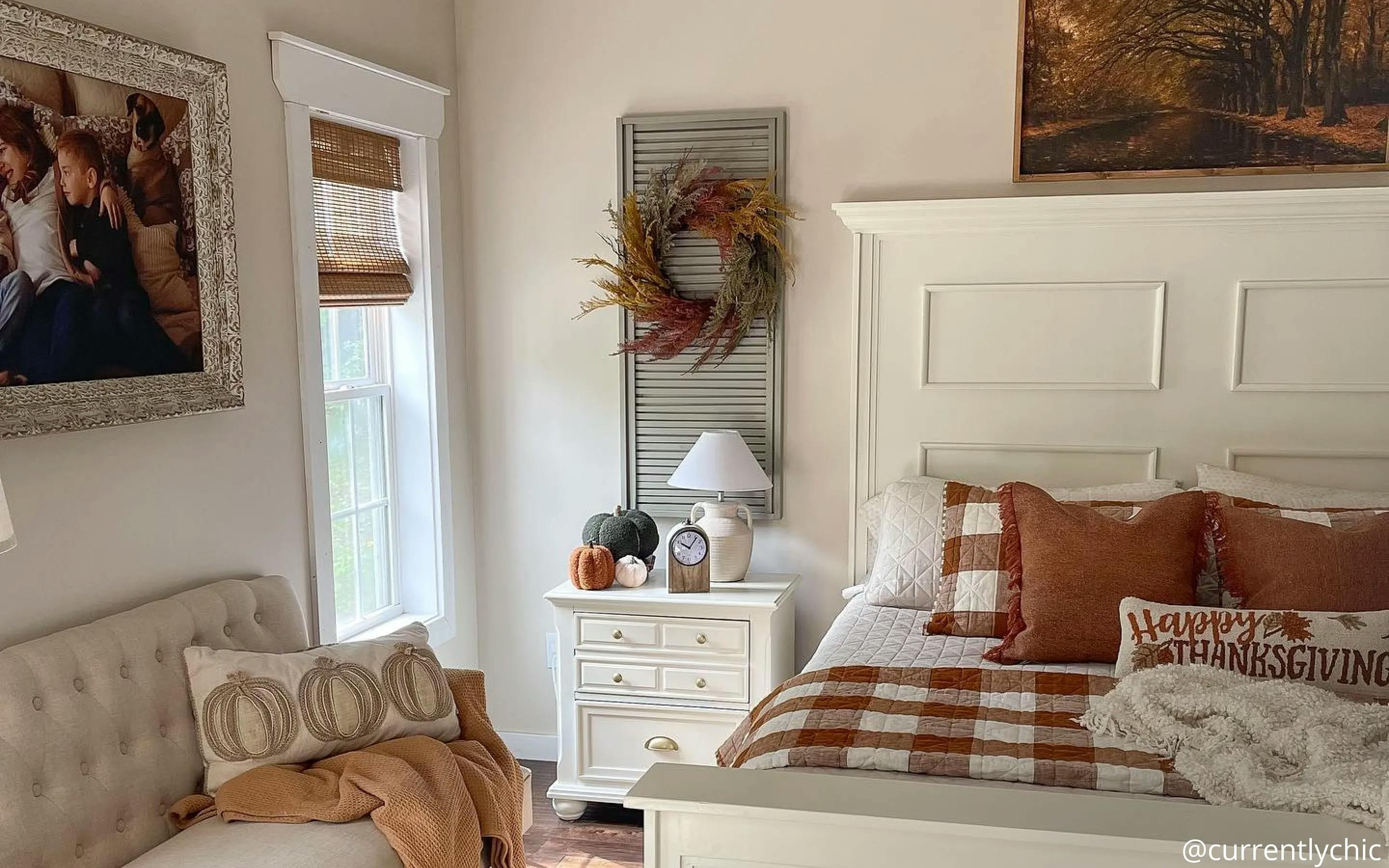 Bedroom with white furniture and orange, brown and gingham bedding. The window features woven wood shades. On the wall to the left of the window is a picture frame. Under the frame is a neutral-colored couch with an orange throw blanket and pumpkin-patterned throw pillow.