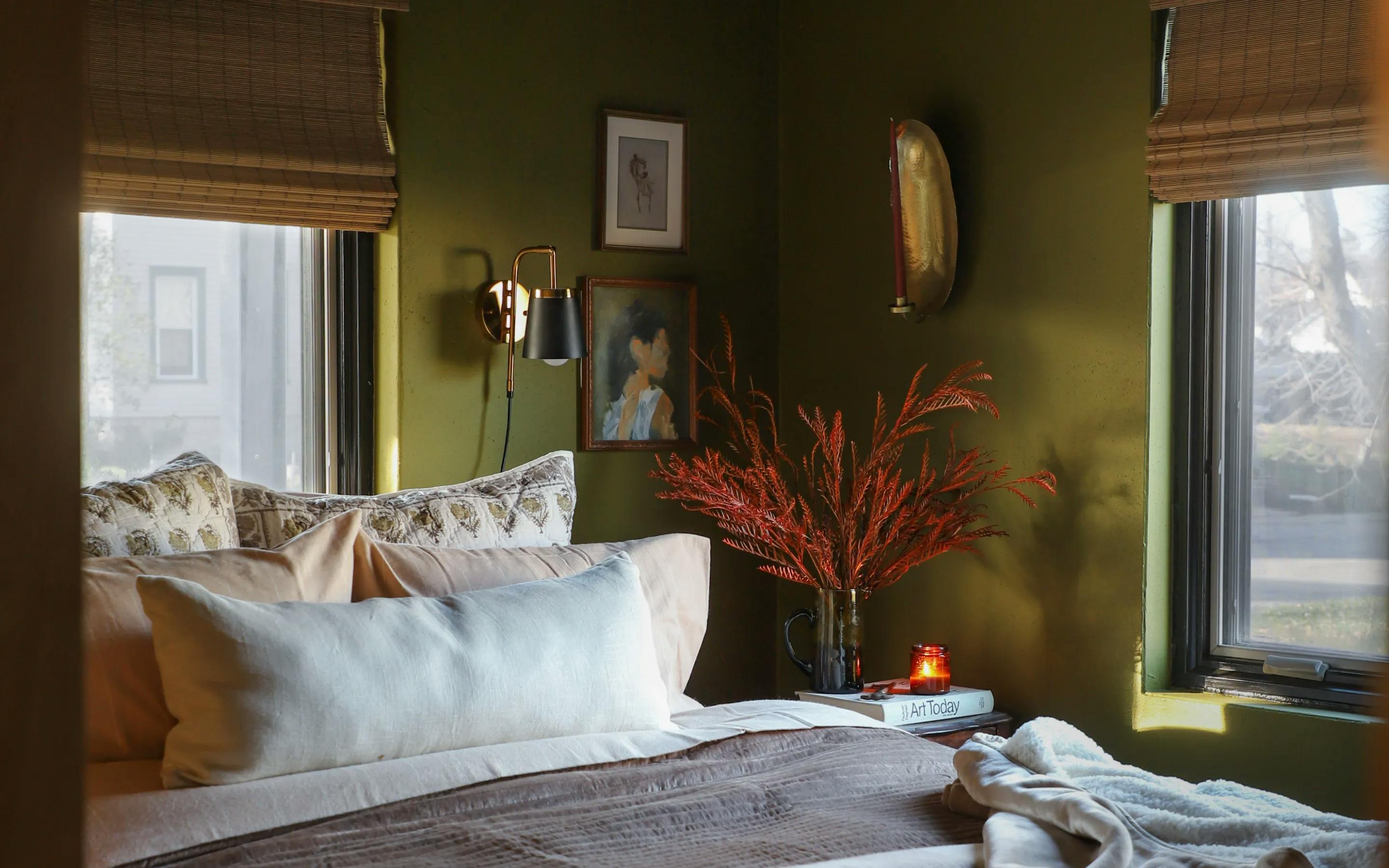 Bedroom with mossy green walls and windows covered with woven wood shades. Against one of the windows is a bed with cream and brown bedding. The side table has a vase with red foliage in it a and a candle on top of books.