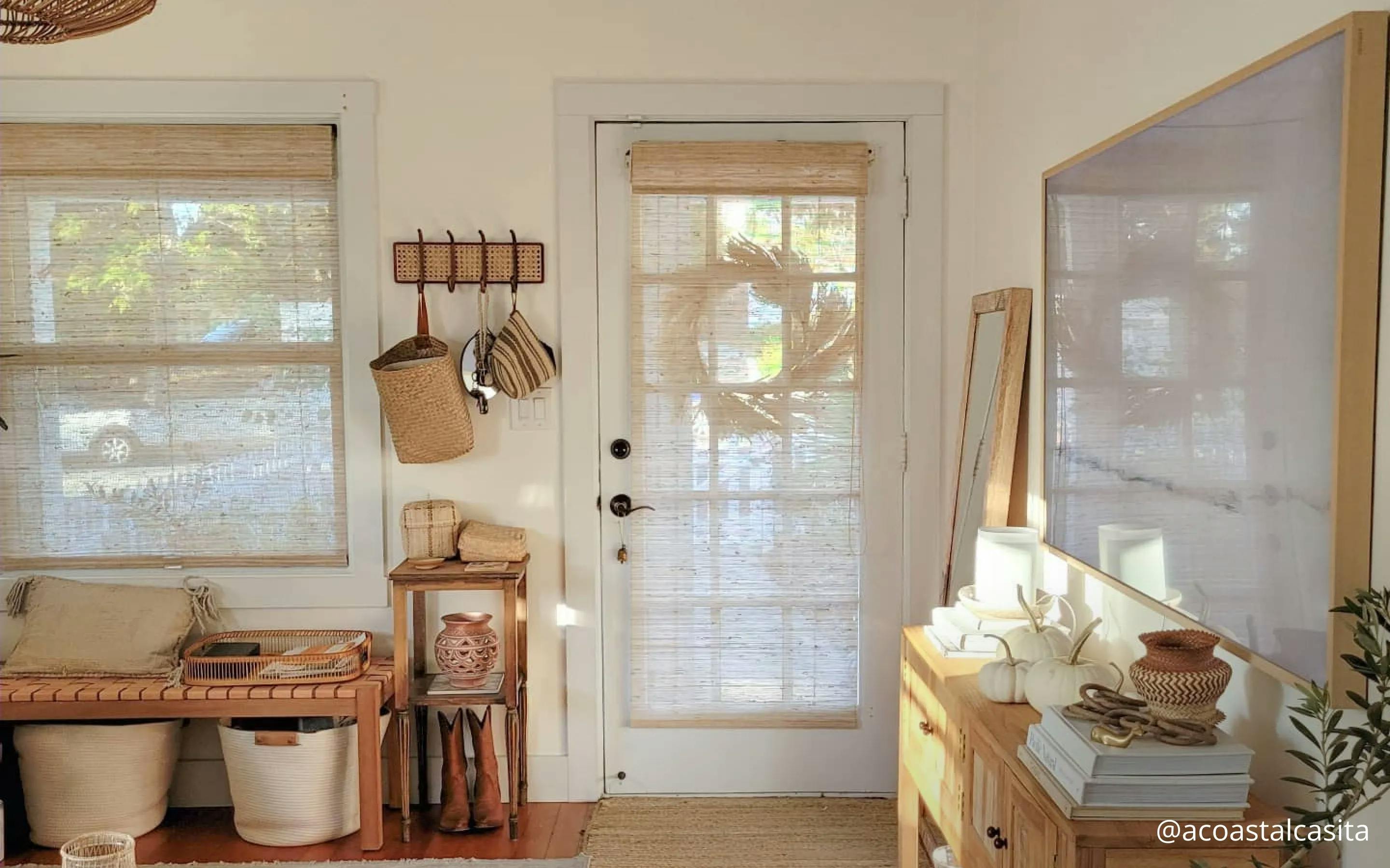 Entryway with a door covered in light woven wood shades. The floormat in front of the door is of the same color. To the left of the door is a bench with baskets, a place to hang coats and a small shoe stand. There's a window above the bench that features matching shades.