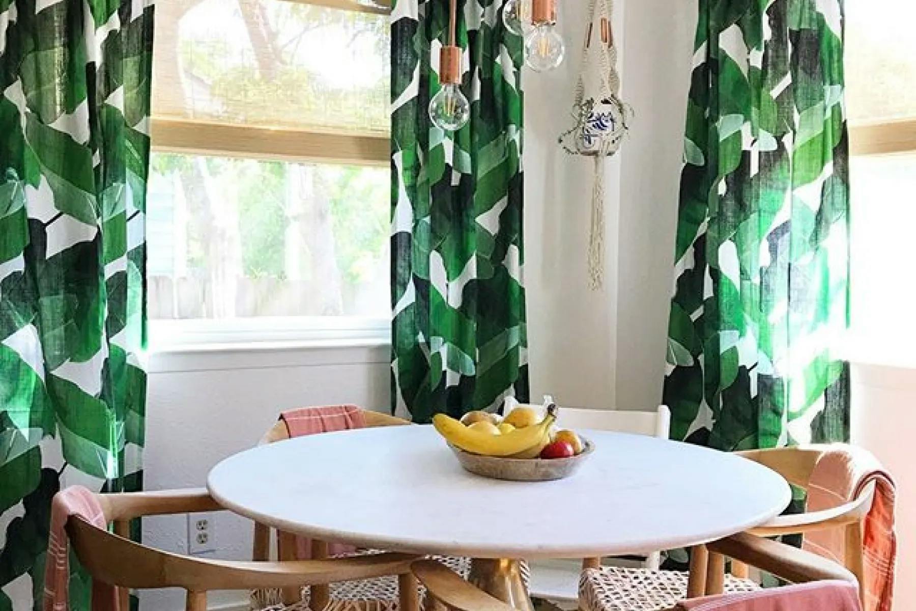 Dining space with light-filtering roller shades layered below bold, leaf-patterned curtains.