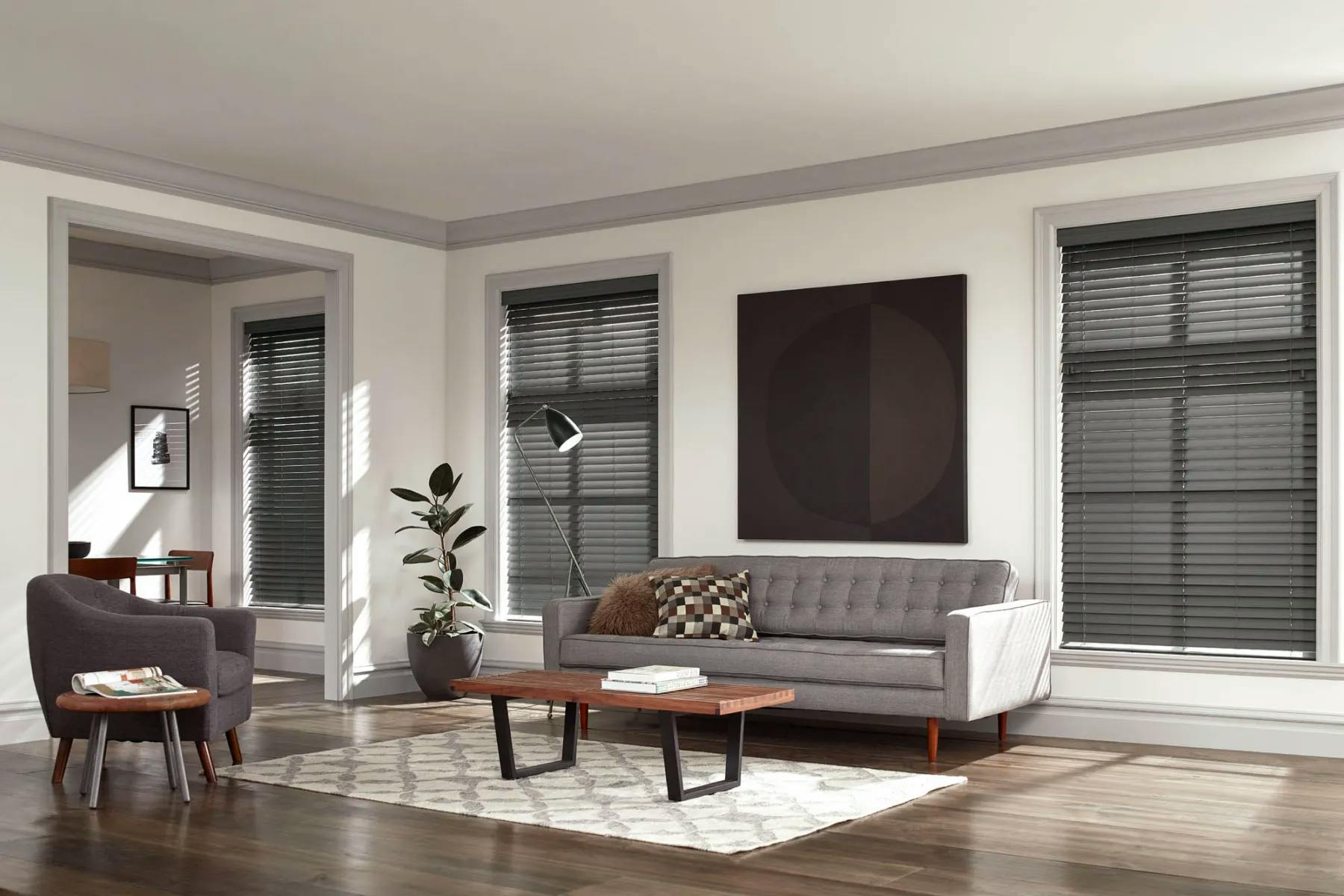 Living room with dark, warm neutrals. Large windows frame a gray couch. Deep gray wood blinds are on th windows.