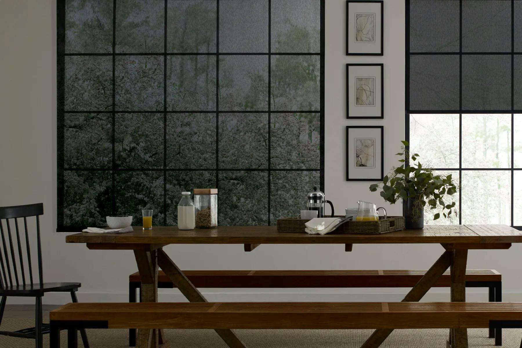 Dining space with a long, wooden table. Large windows are on the back wall and are covered in black, light-filtering solar shades.