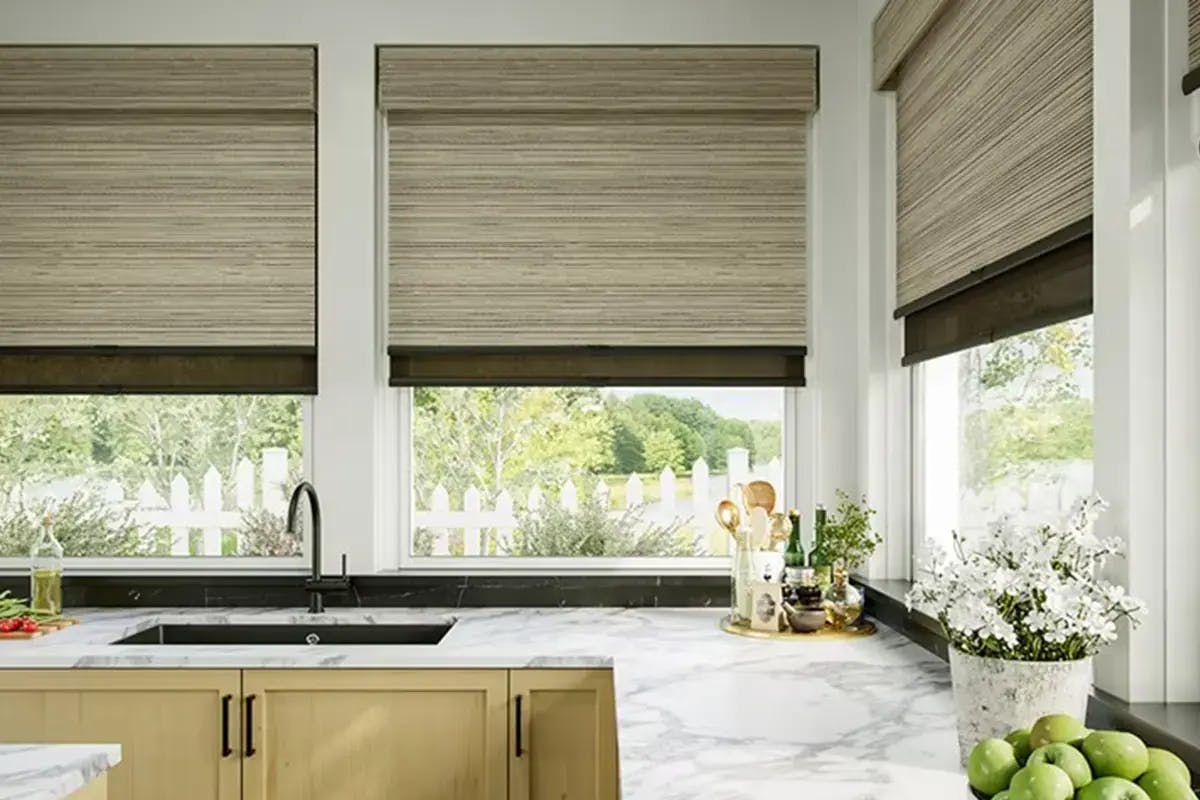 Kitchen with roller shades on three windows above a sink and marble countertop.