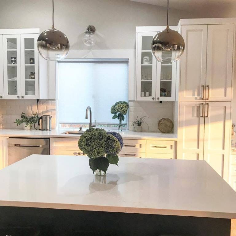 white kitchen with metallic globe pendant lights, large island and flat white roller shade behind sink