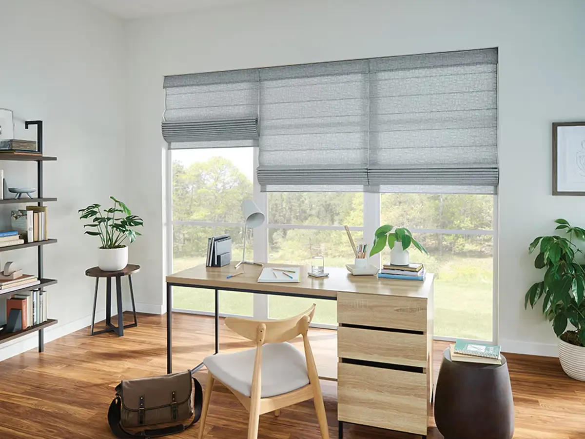 A home office with three light grey textured Roman shades installed on a large window.