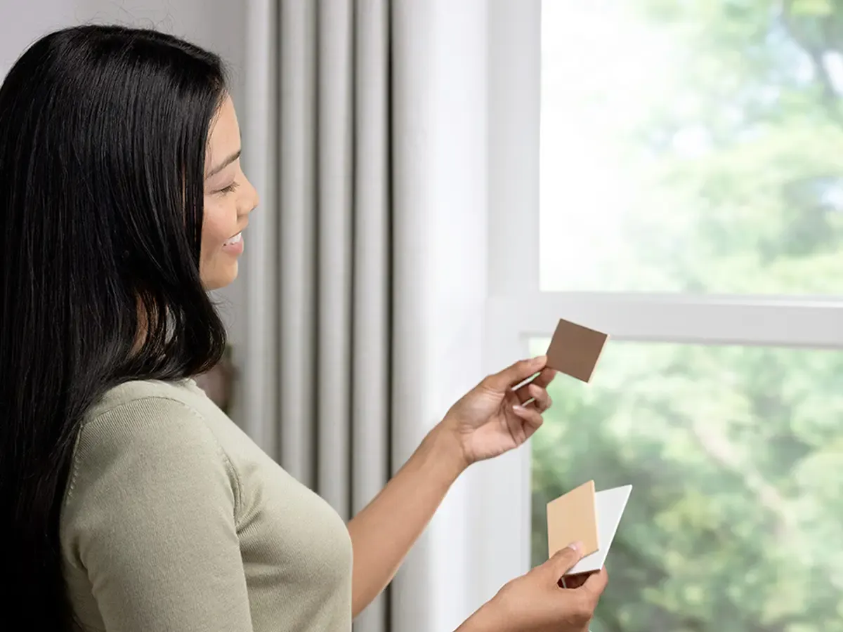 A woman standing by a window holding up various samples.
