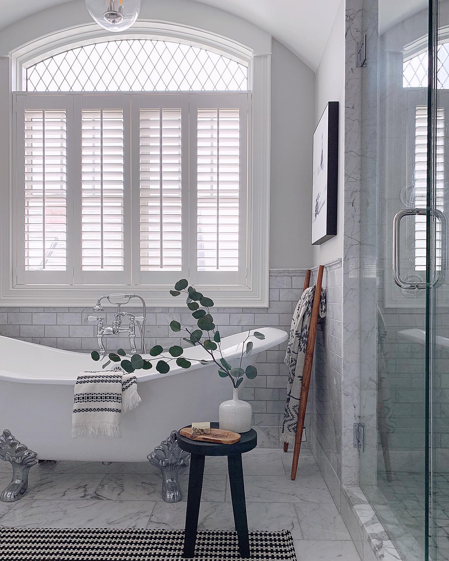 All-white bathroom with white Bllinds.com shutters and a claw foot tub. There is a small black stool beside the tub with a plant in a white vase.
