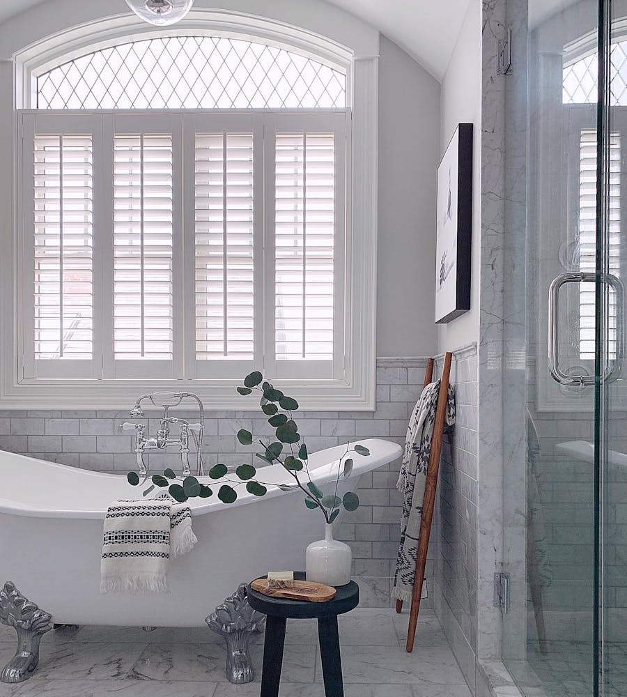 All-white bathroom with white Bllinds.com shutters and a claw foot tub. There is a small black stool beside the tub with a plant in a white vase.