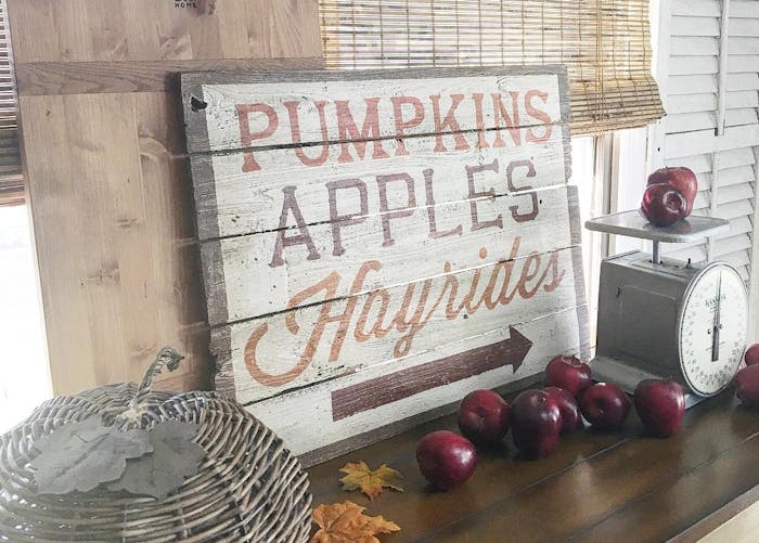 A table decorated for fall with a decorative fall sign, straw pimpkin, and red apples arranged on a table