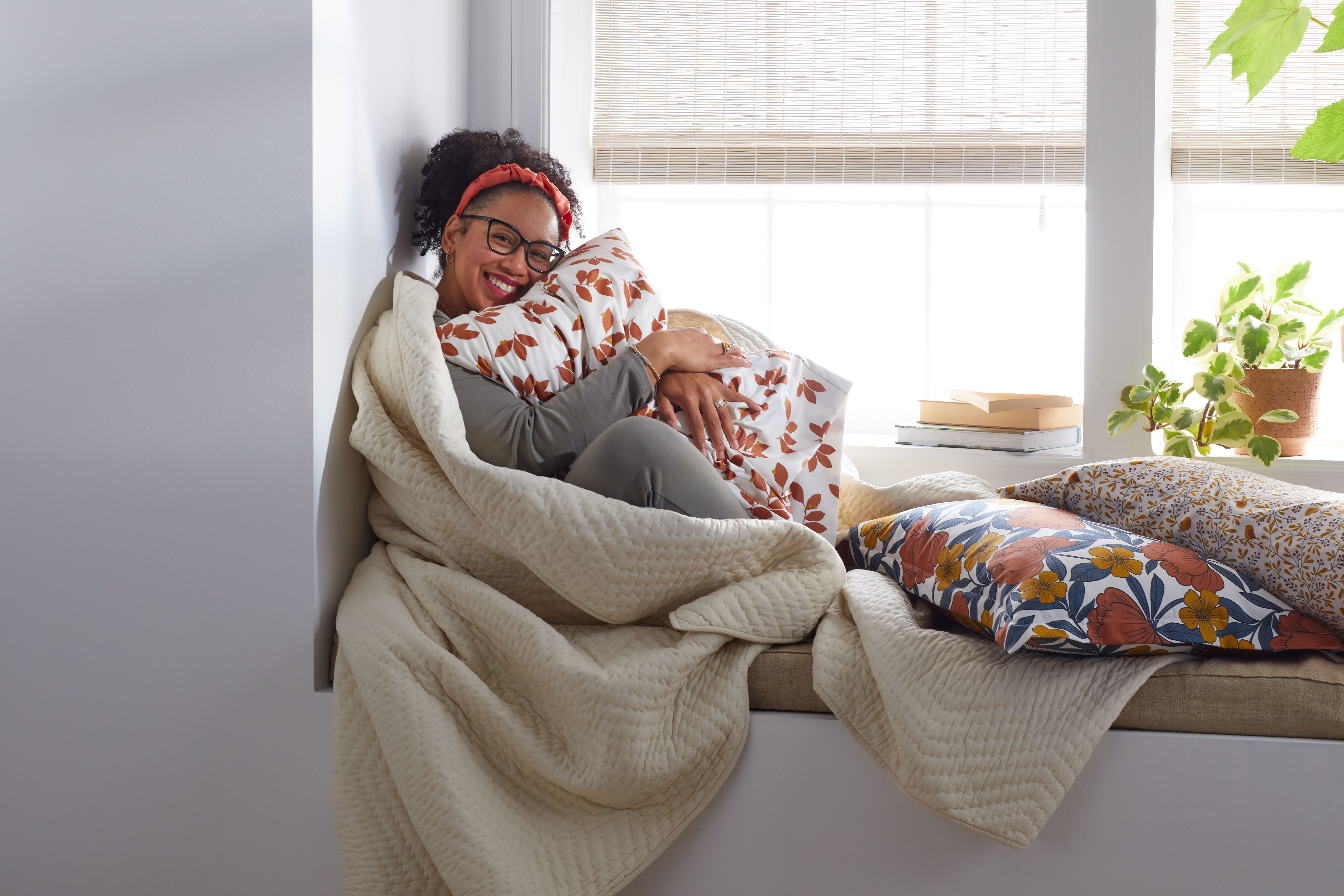 A woman smiling at home sitting in front of Blinds.com Woven Wood Shades in Rustic Jute.