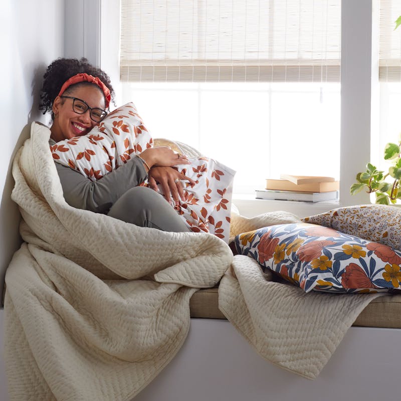 A woman smiling at home sitting in front of Blinds.com Woven Wood Shades in Rustic Jute.