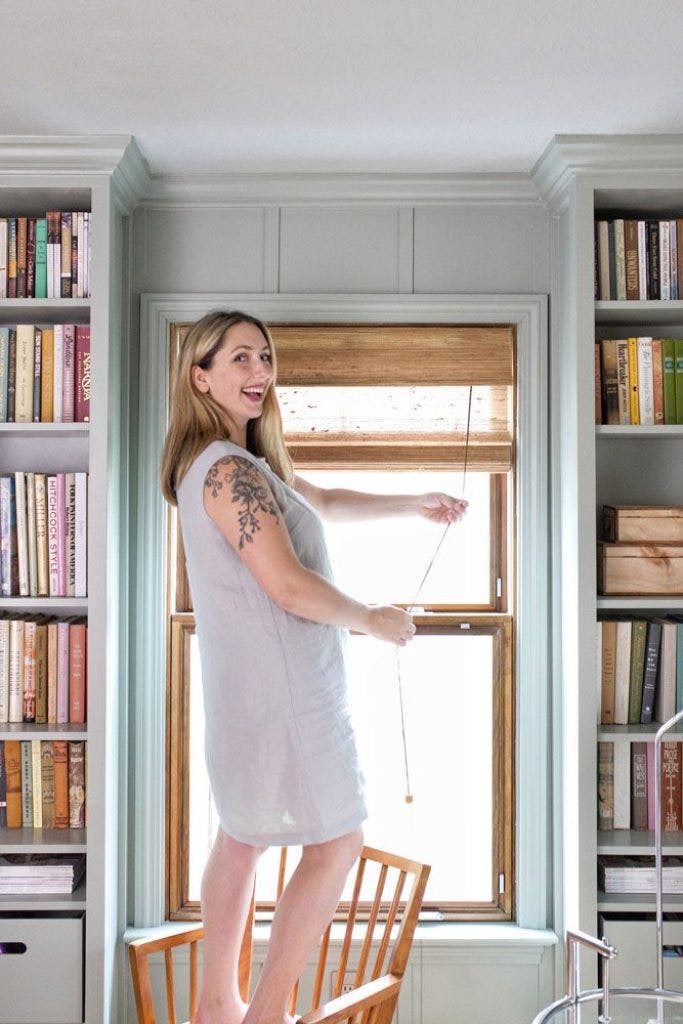 Mandi makes blogger standing on chair adjusting bamboo blinds in home office with built in bookcases