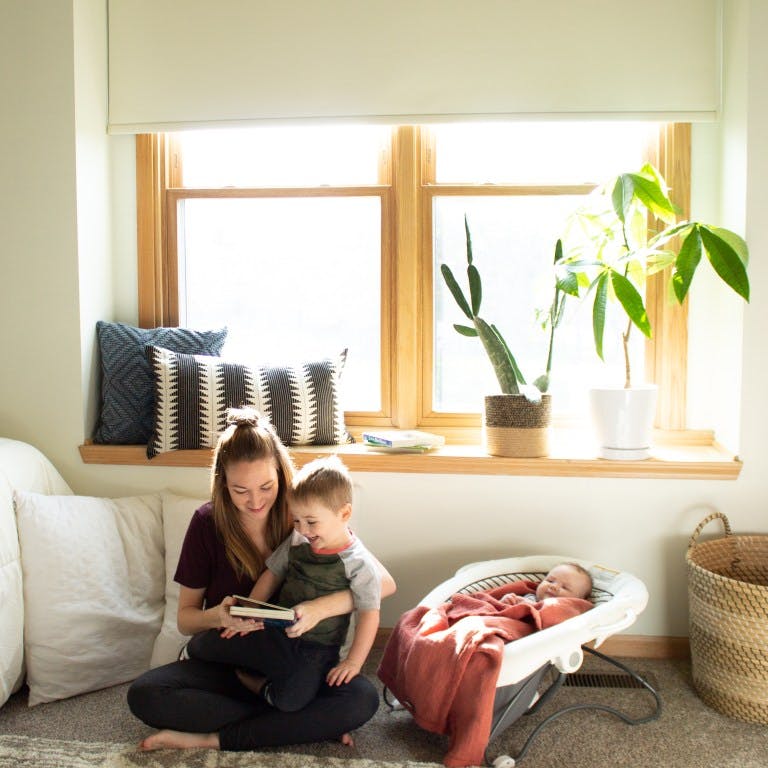 mom reading book to toddler and infant in eclectic nursery under window with roller shade