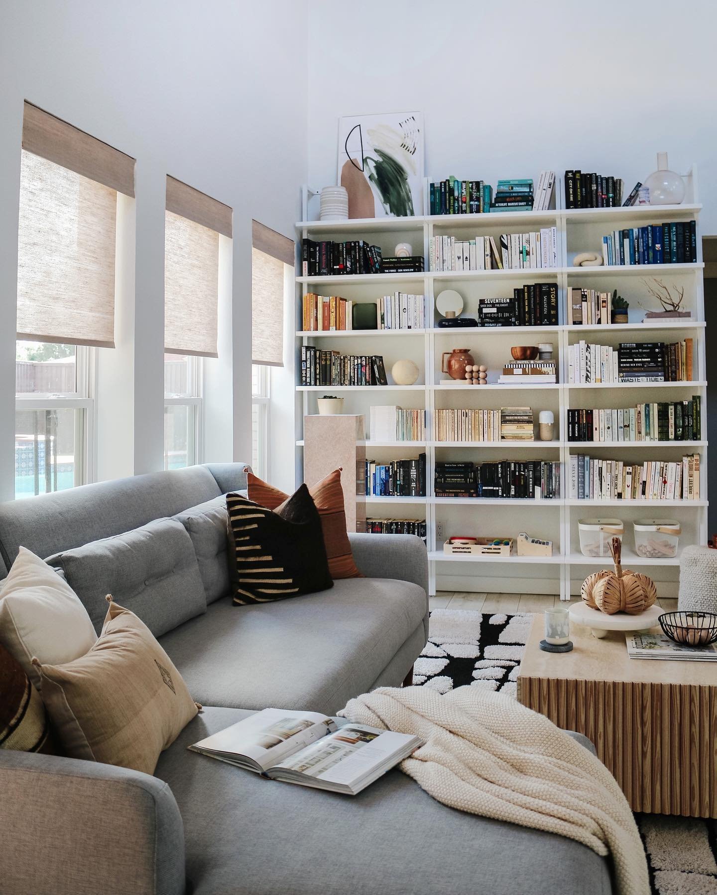 A living room decorated with fall home decor. A grey couch in front of windows with woven roller shades from Blinds.com. There is a bookshelf filled with books next to the sofa.