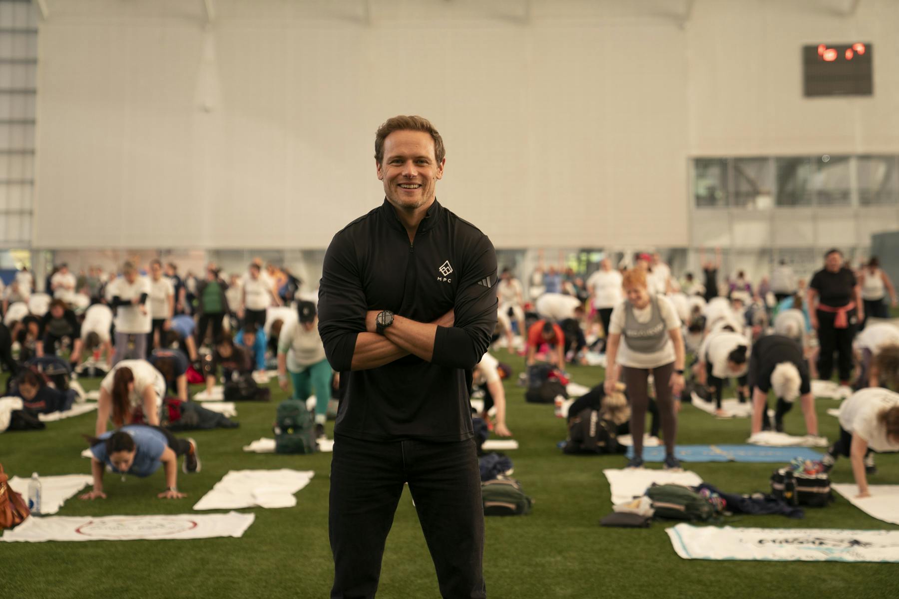 Sam Heughan smiling in athletic wear in front of a group of people exercising.
