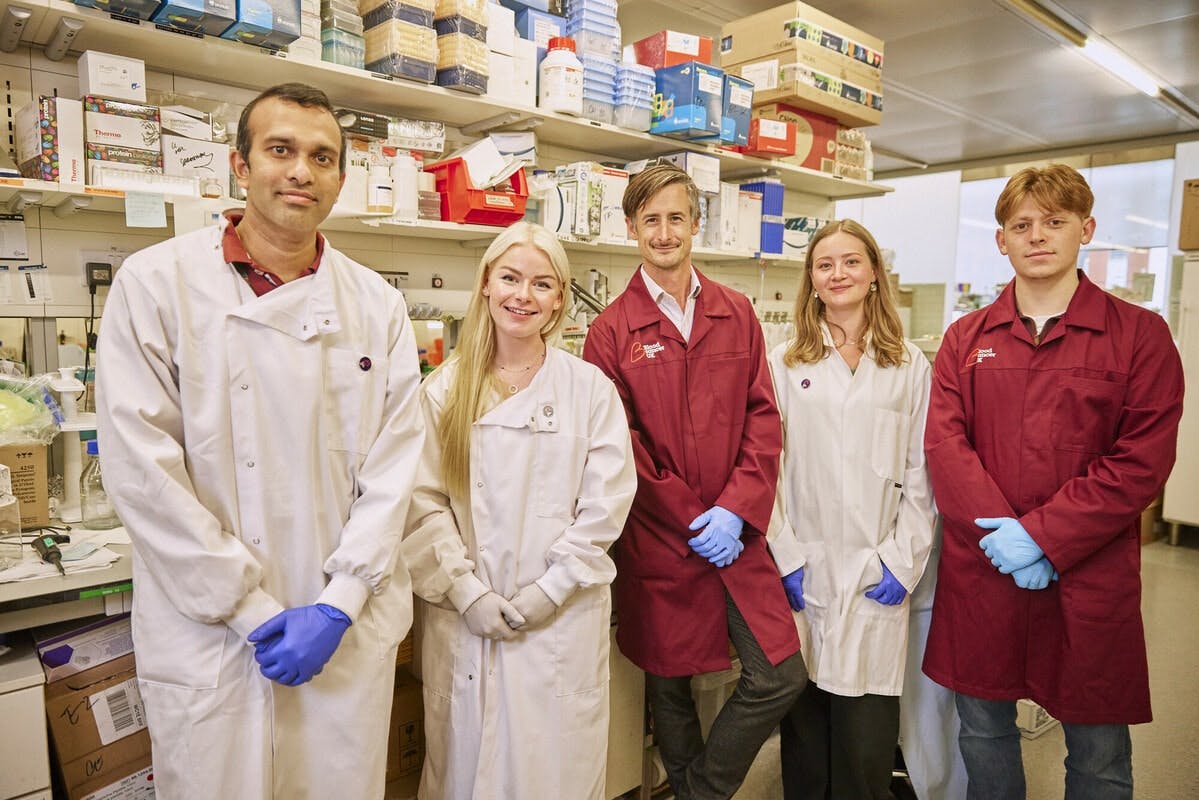 Five researchers standing together in front of shelves with boxes. All wearing white or burgundy lab coats