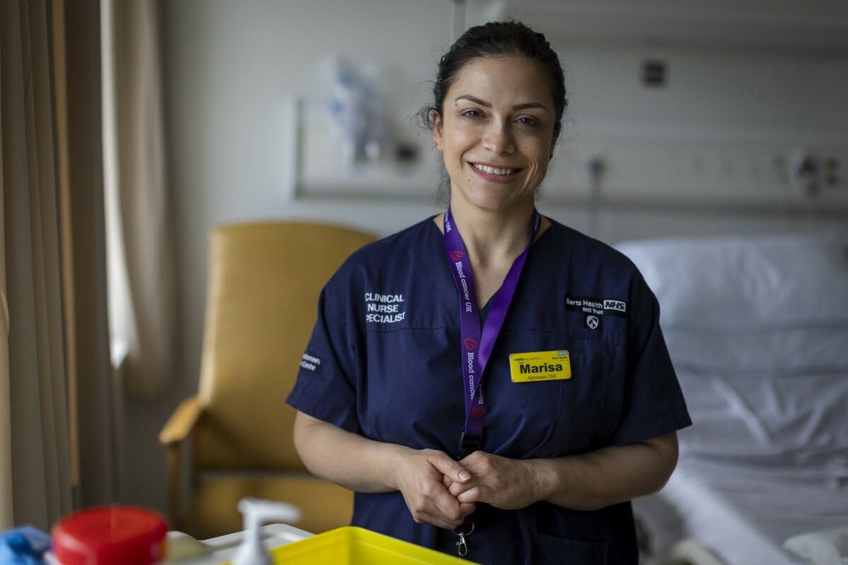 A clinical nurse specialist standing next to a hospital bed wearing her uniform and a Blood Cancer UK lanyard