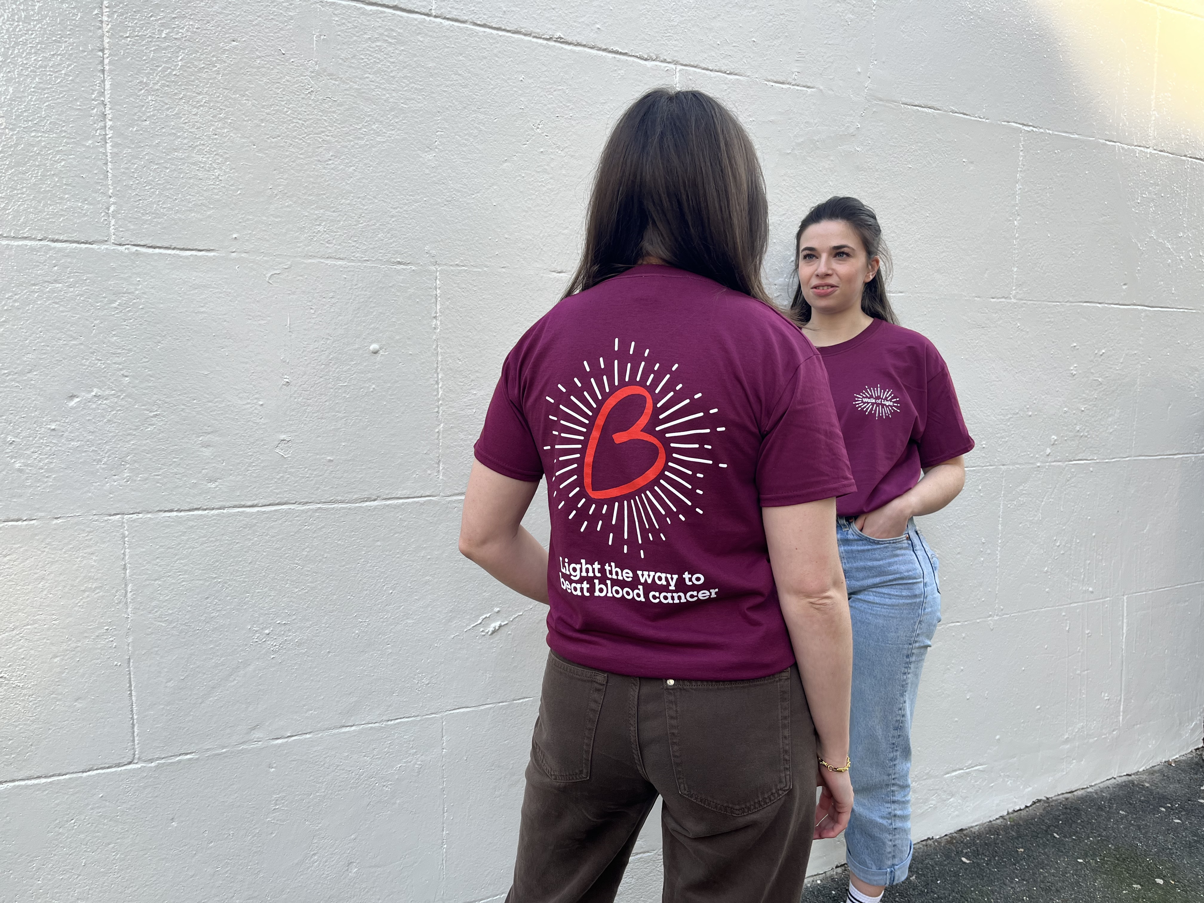 Two people facing each other wearing the Walk of Light t shirt. The back of the shirt has a large red B heart with white lines coming off it