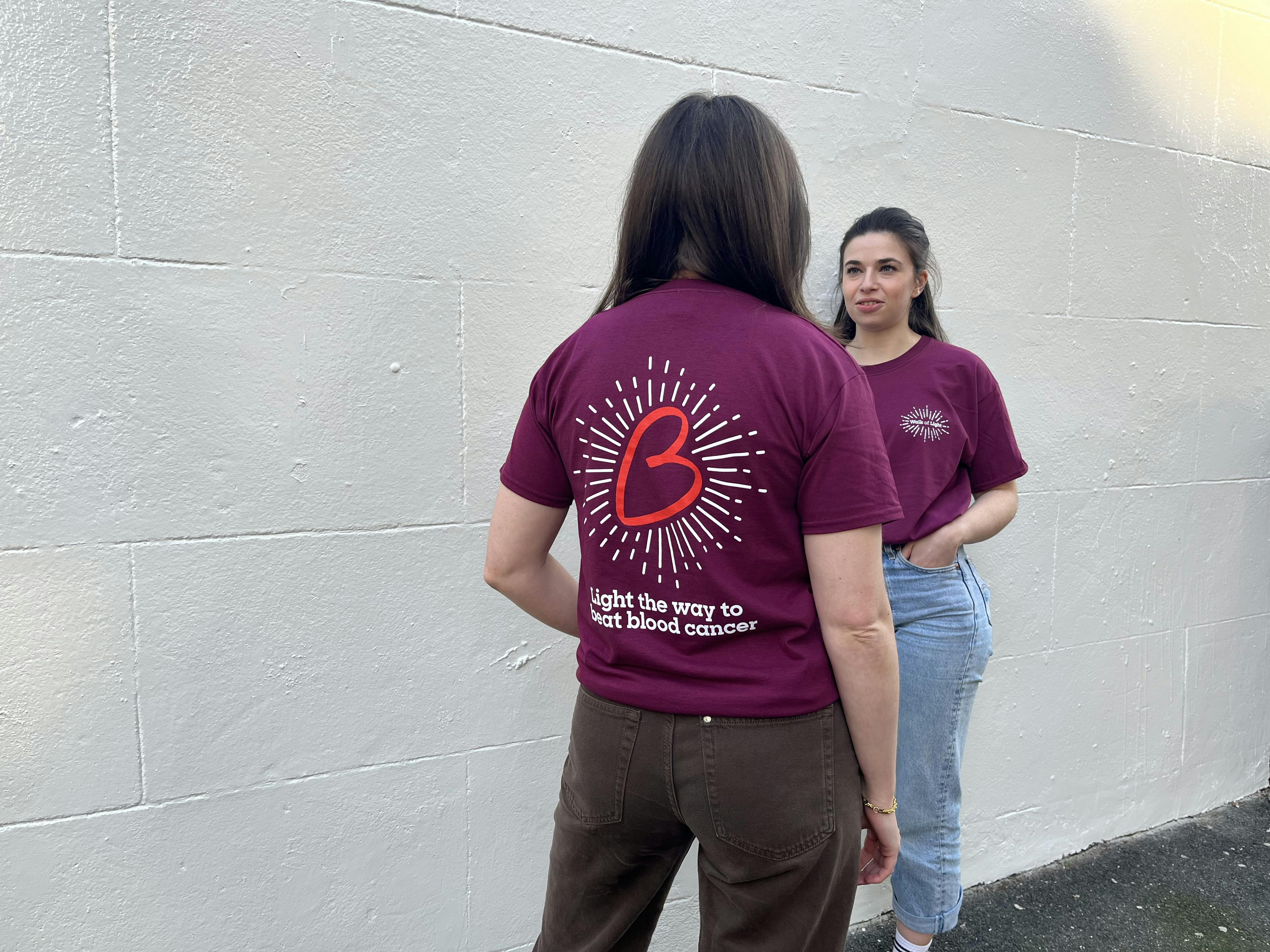 Two people facing each other wearing the Walk of Light t shirt. The back of the shirt has a large red B heart with white lines coming off it