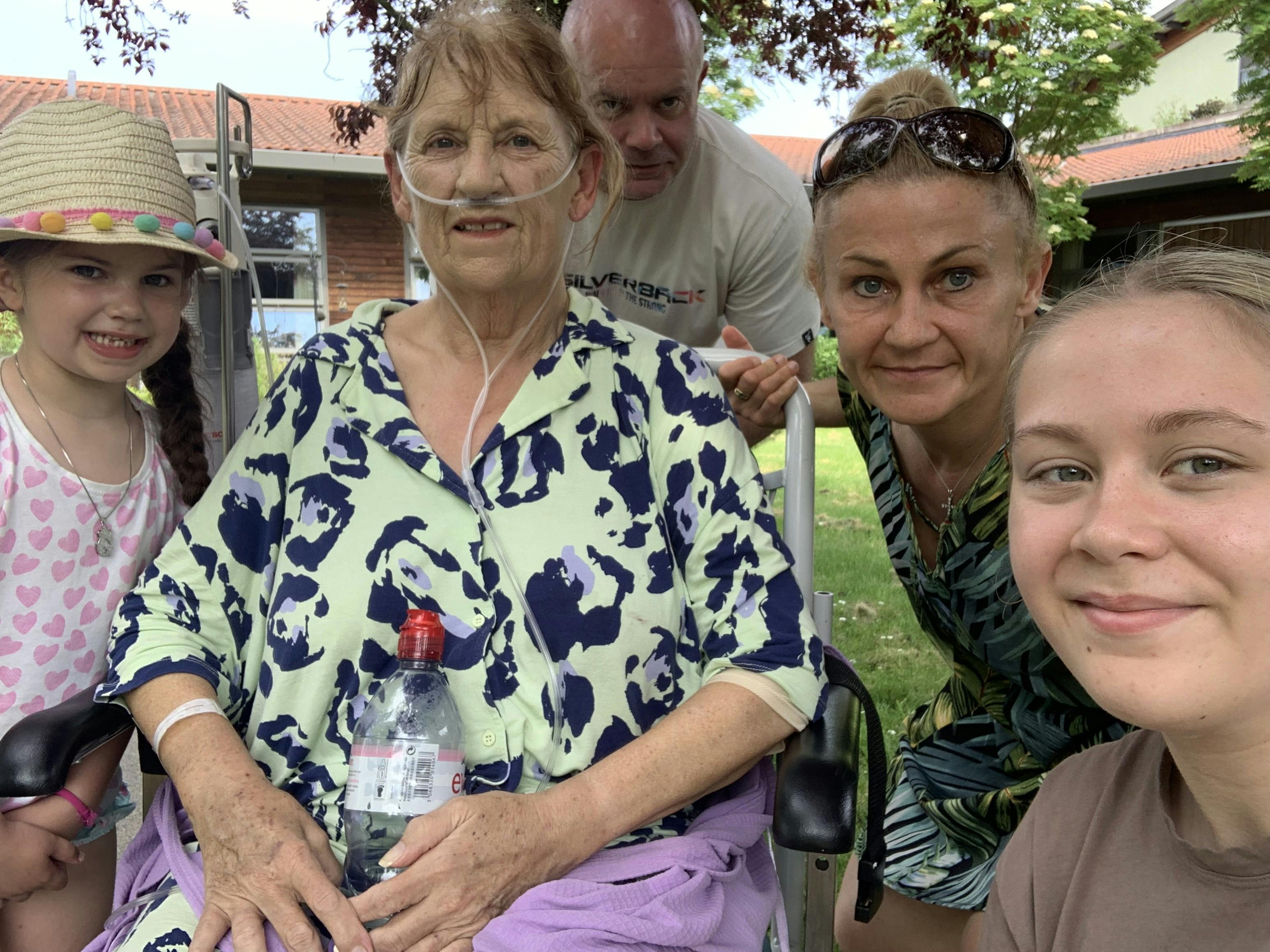 Elderly person in a wheelchair outdoors, wearing a patterned top and holding a water bottle, surrounded by family members.