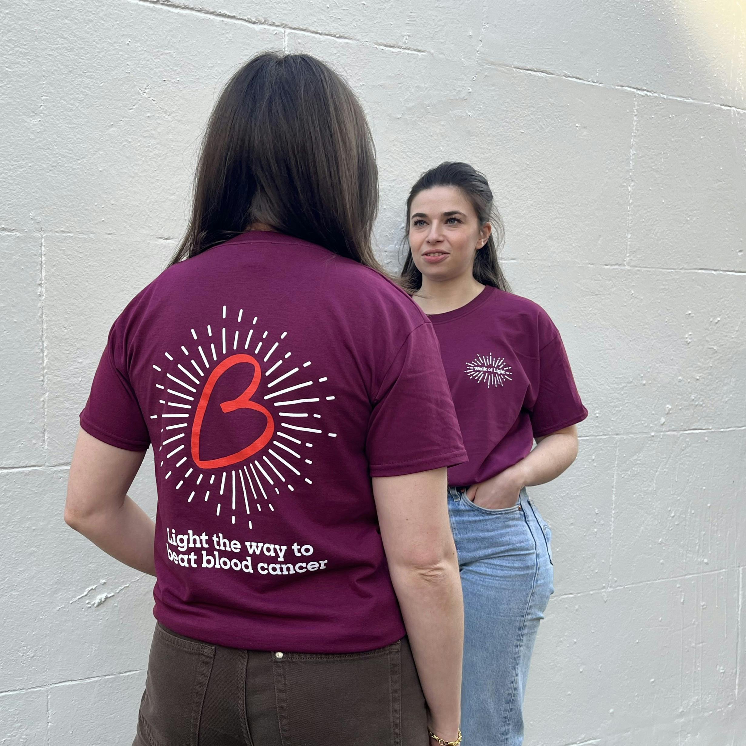 Two people facing each other wearing the Walk of Light t shirt. The back of the shirt has a large red B heart with white lines coming off it