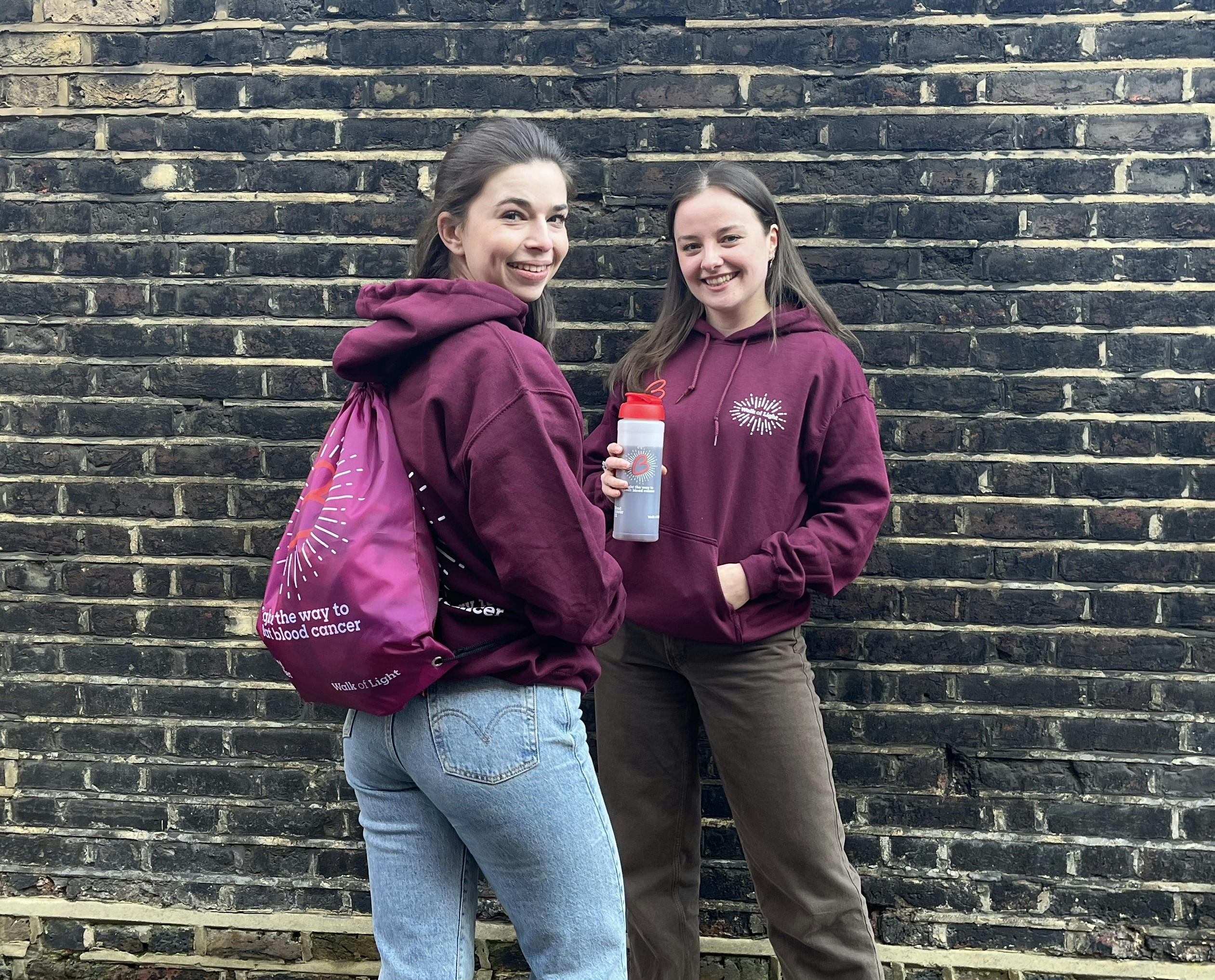 Two people wearing matching maroon hoodies, one holding a water bottle and the other carrying a maroon drawstring bag, standing in front of a brick wall.