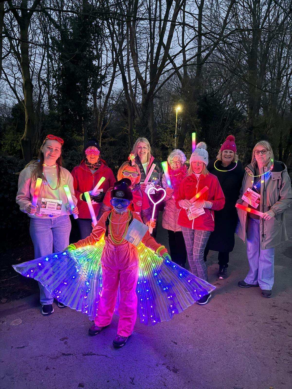 Group of people outdoors at dusk holding glowing sticks, with one person in front wearing illuminated rainbow wings.