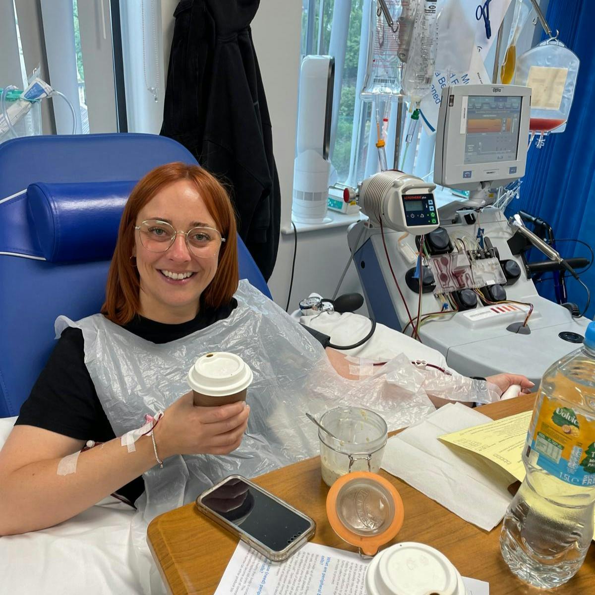 Katie in a hospital chair with medical equipment nearby, holding a takeaway coffee cup and surrounded by treatment items.