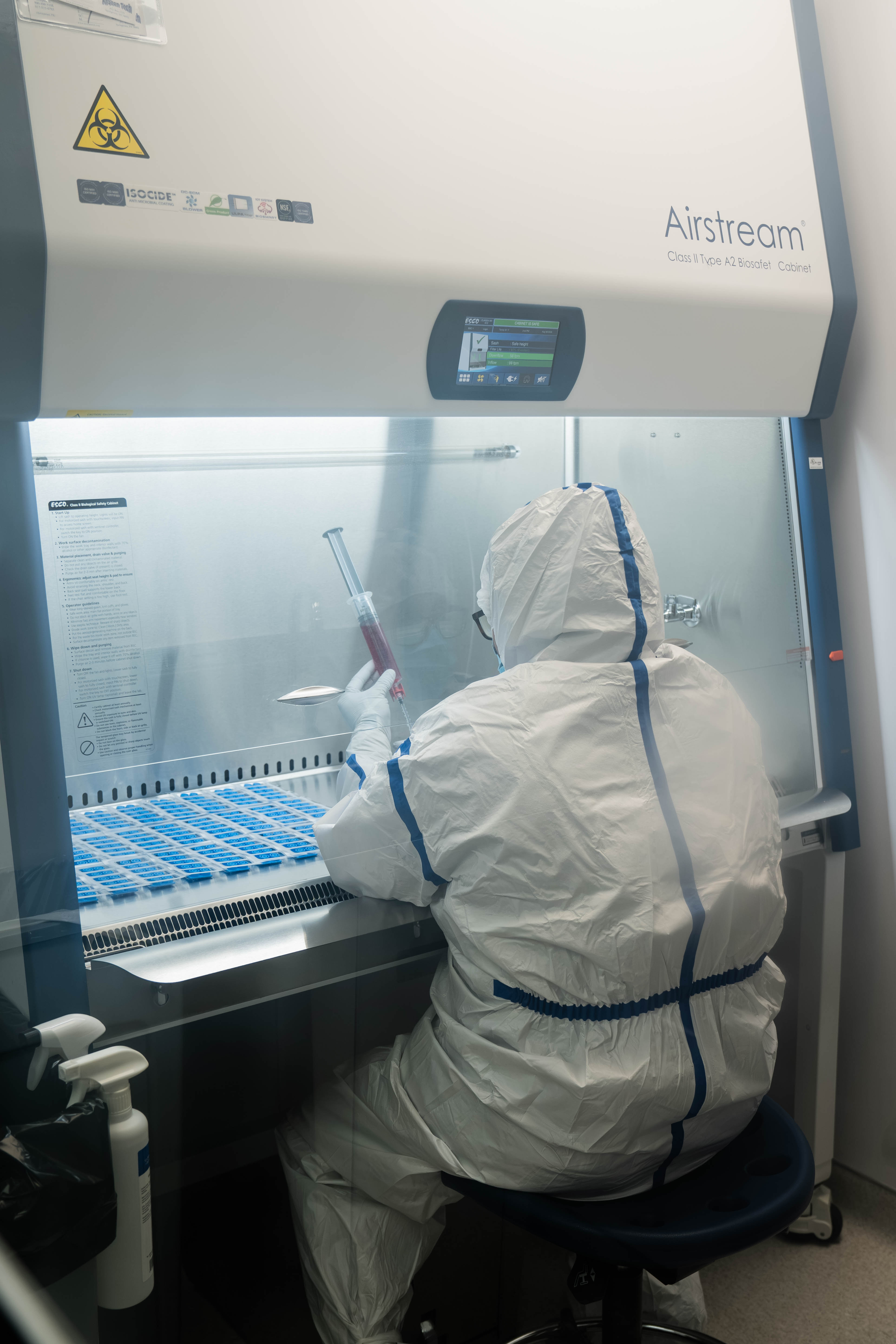A pharmacist in full protective gear compounding a medication inside a sterile biosafety cabinet.