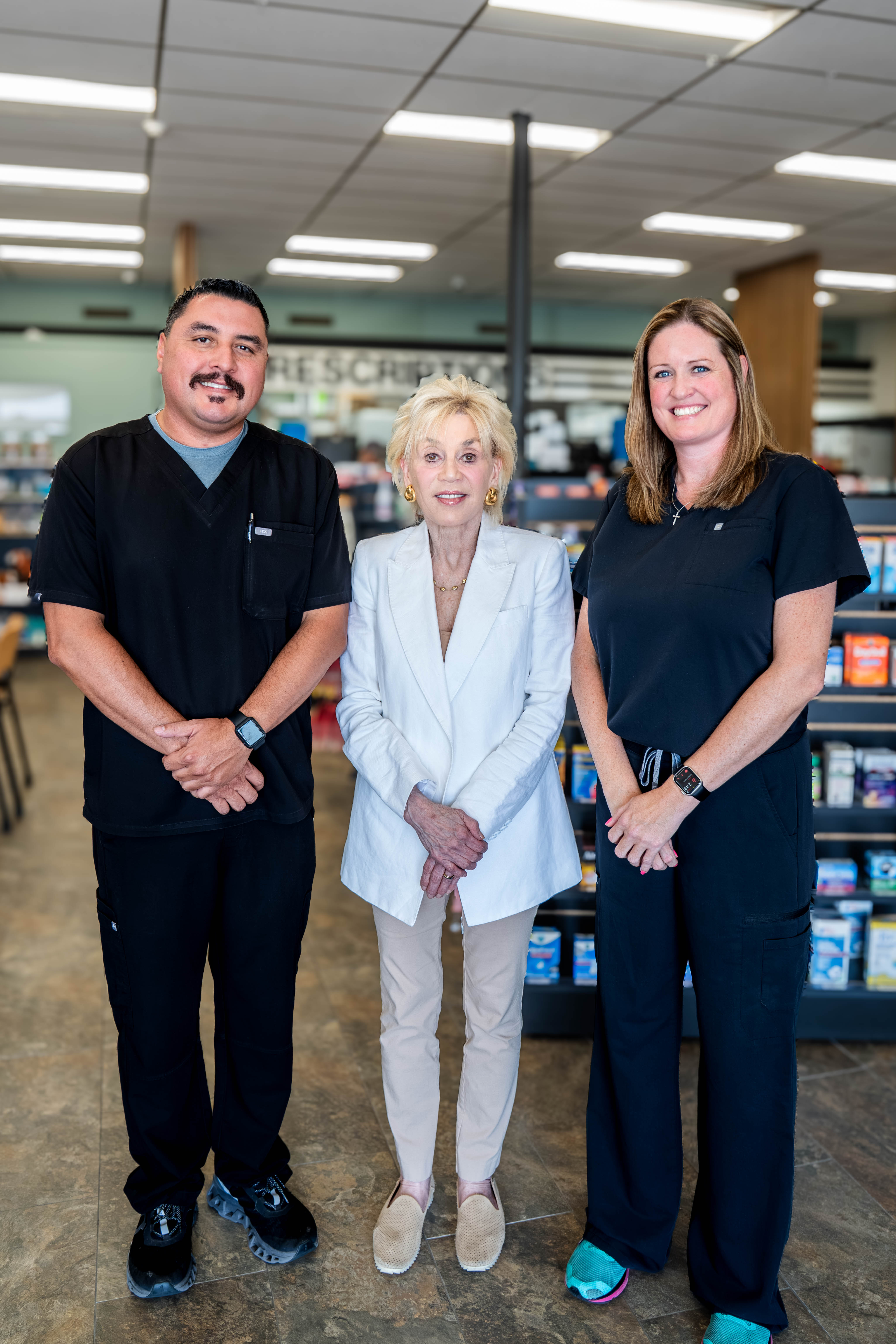 Three members of the Perrone Pharmacy team including Paula Perrone standing together in the pharmacy aisle, two pharmacists in black scrubs on either side of a woman in a white suit.