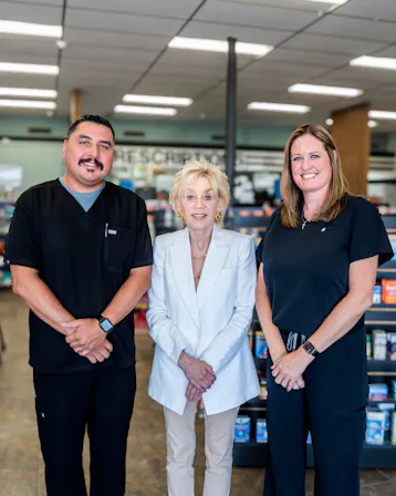 Three members of the Perrone Pharmacy team including Paula Perrone standing together in the pharmacy aisle, two pharmacists in black scrubs on either side of a woman in a white suit.