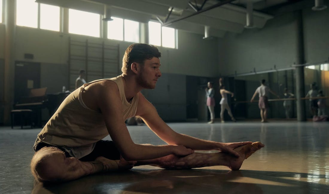 An image of a man sitting on the floor in a dance studio, mirrors and ballet barres can be seen in the background.