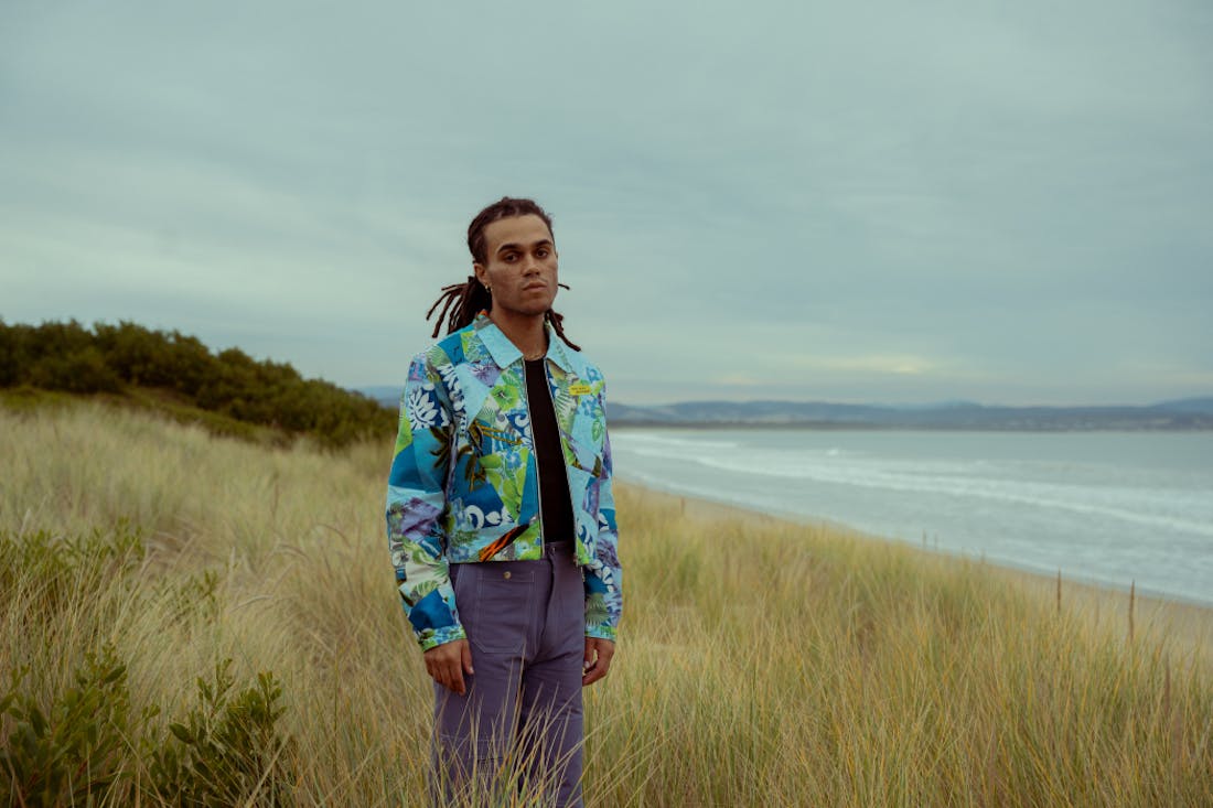 An image of a man standing in long grass beside the beach, he is wearing a vibrant jacket.