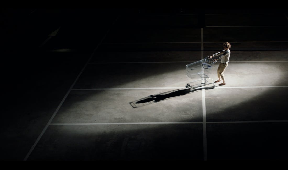 An image of a person illuminated by a spotlight, pushing a shopping trolley through a carpark at night.