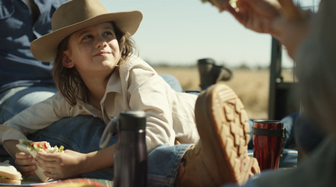 An image of a young girl wearing a wide brim hat and work shirt. She is sitting outside with two people eating a sandwich.