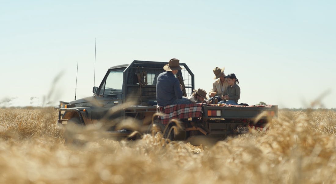 An image showing a man, a woman and two children sitting on the back of a ute in a field of wheat on a sunny day.