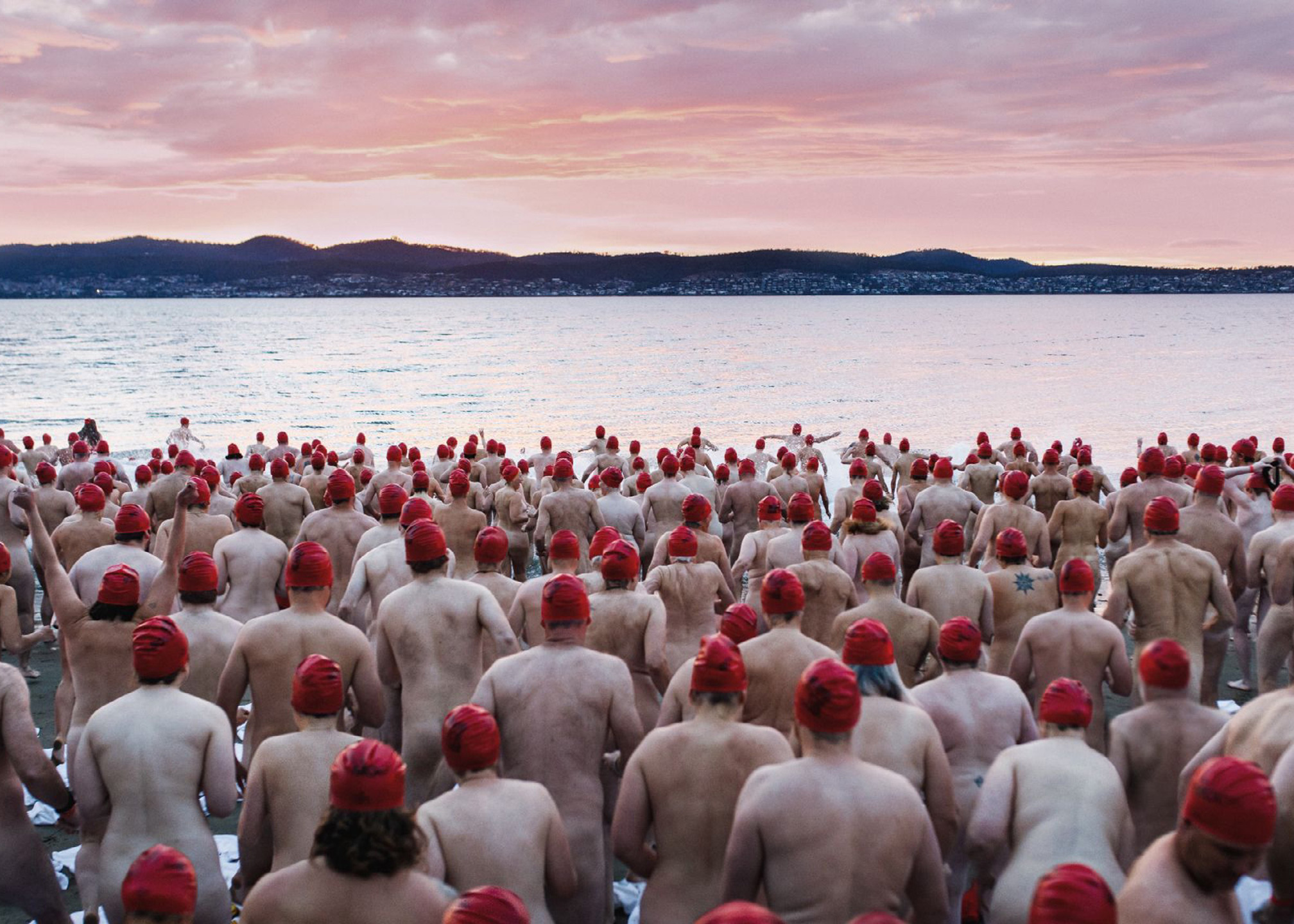 An image of a large group of people in red swimming caps, wading into a serene lake at sunrise to go skinny dipping.