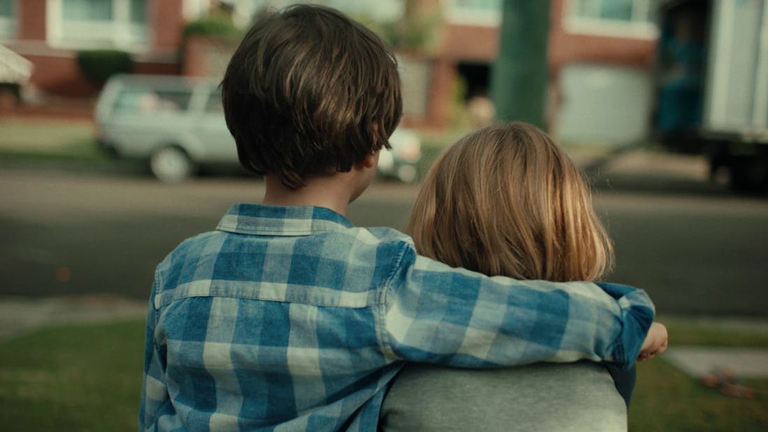 An image of two kids embracing each other, looking out over a street to an apartment building.