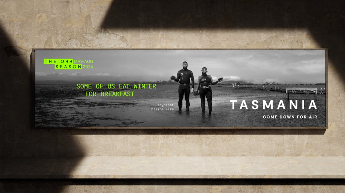 Black and white billboard of two divers holding sea urchins on a beach with green text reading 'The Off Season May-Aug 2024, Some Of Us Eat Winter For Breakfast, Tasmania Come Down For Air'