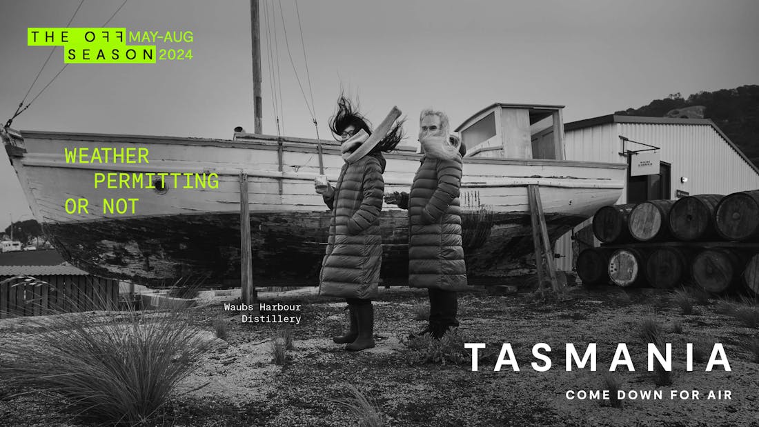 Black and white billboard of a couple in long winter coats in front of a boat with green text reading 'The Off Season May-Aug 2024, Weather Permitting or Not, Tasmania Come Down For Air'
