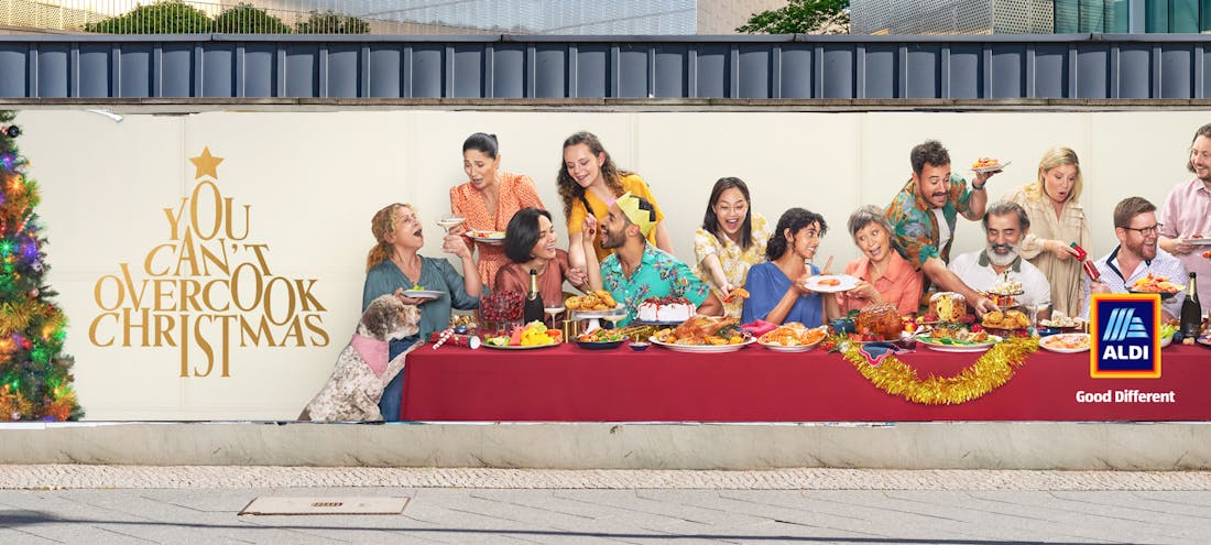 A billboard advertisement for ALDI showing a large group of people enjoying a feast on a long table. The tagline reads: "You can't overcook Christmas".