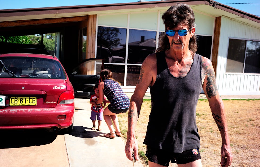 An image of a man wearing a blue singlet and a pair of sunglasses walking towards the camera. A young woman picks up a small child in the background. 